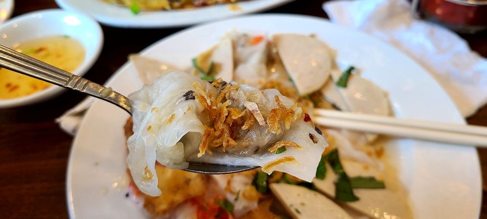 A freshly steamed banh cuon rice roll lifted on a spoon, topped with fried shallots and herbs